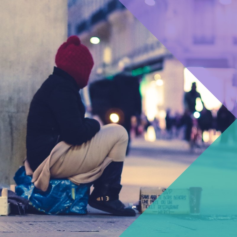 A photo of an unhoused person sitting on a box in the street with a cardboard sign